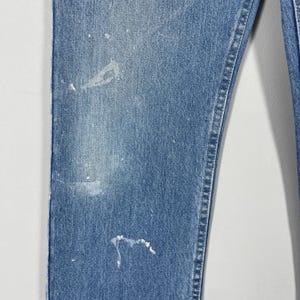 以下が含まれることがあります： 白いペンキの飛沫が付いた青いデニムジーンズのクローズアップ。ジーンズは、クラシックな5ポケットのデザインとストレートレッグカットです。デニムは色あせたウォッシュ加工が施されています。