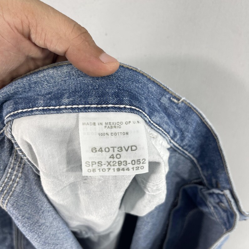 K&ouml;nnte beinhalten: Ein blaues Jeans-Etikett mit wei&szlig;en N&auml;hten, auf dem "MADE IN MEXICO OF U.S. FABRIC 100% COTTON 640T3VD 40 SPS-X293-052 051071944120" steht.