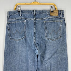 May include: Light blue denim Wrangler jeans, featuring the classic W-shaped stitching on the back pockets. The jeans are hanging on a yellow hanger. The Wrangler label is visible on the back.