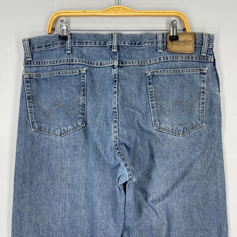 May include: Light blue denim Wrangler jeans, featuring the classic W-shaped stitching on the back pockets. The jeans are hanging on a yellow hanger. The Wrangler label is visible on the back.