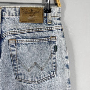 Pode incluir: Jeans de denim azul claro com um remendo de couro marrom que diz "Wrangler" e um bolso traseiro com um logotipo "W" costurado.