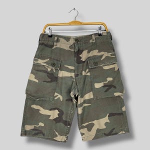 K&ouml;nnte beinhalten: Tarnfarbene Cargo-Shorts in Gr&uuml;n-, Braun- und Beiget&ouml;nen, h&auml;ngen an einem Holzb&uuml;gel. Die Shorts haben mehrere Taschen und einen Knopfverschluss. Die Shorts sind etwa 51 cm lang.
