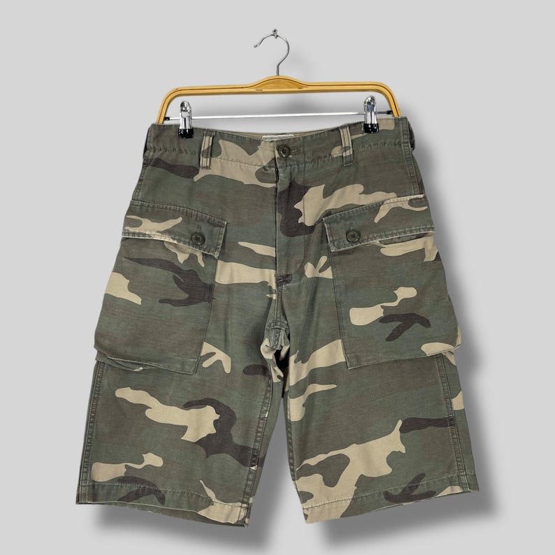 K&ouml;nnte beinhalten: Tarnfarbene Cargo-Shorts in Gr&uuml;n-, Braun- und Beiget&ouml;nen, h&auml;ngen an einem Holzb&uuml;gel. Die Shorts haben mehrere Taschen und einen Knopfverschluss. Die Shorts sind etwa 51 cm lang.