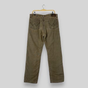 May include: Olive green denim jeans with the Wrangler logo on the back pockets. The jeans are hanging on a gold-colored hanger. The jeans have a straight leg cut and a brown leather patch on the back.