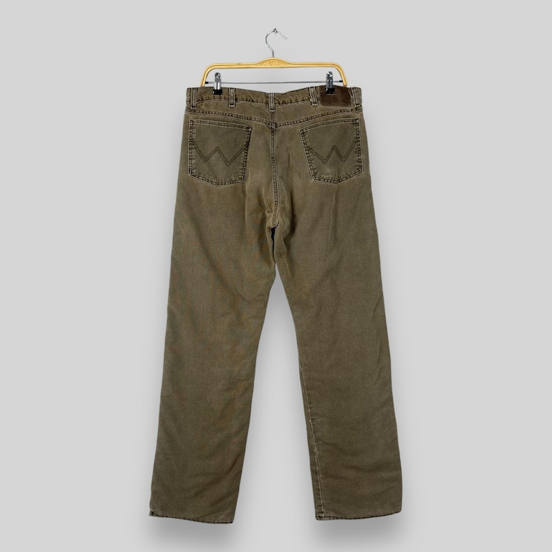 May include: Olive green denim jeans with the Wrangler logo on the back pockets. The jeans are hanging on a gold-colored hanger. The jeans have a straight leg cut and a brown leather patch on the back.