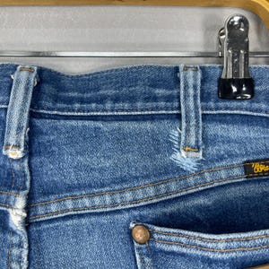 Pode incluir: Close-up de cal&ccedil;as de ganga azuis com um aspeto desgastado e envelhecido. As cal&ccedil;as de ganga t&ecirc;m um design cl&aacute;ssico de cinco bolsos, costuras vis&iacute;veis e um bot&atilde;o de metal. A etiqueta da marca Wrangler &eacute; vis&iacute;vel no bolso de tr&aacute;s.