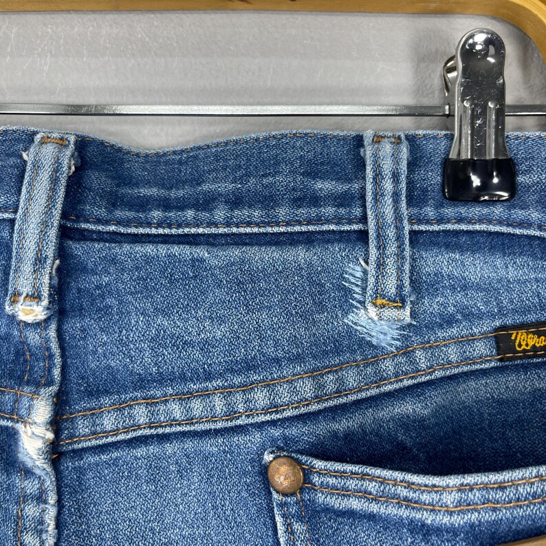 Pode incluir: Close-up de cal&ccedil;as de ganga azuis com um aspeto desgastado e envelhecido. As cal&ccedil;as de ganga t&ecirc;m um design cl&aacute;ssico de cinco bolsos, costuras vis&iacute;veis e um bot&atilde;o de metal. A etiqueta da marca Wrangler &eacute; vis&iacute;vel no bolso de tr&aacute;s.