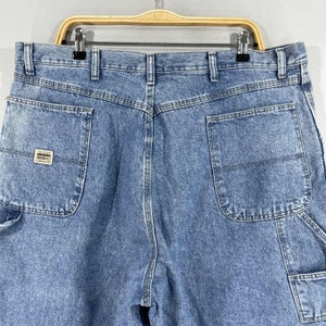 K&ouml;nnte beinhalten: Eine blaue Jeanshose im Carpenter-Stil mit einer Ges&auml;&szlig;tasche, auf der ein wei&szlig;er Etikett mit dem Text "Los Angeles Originals" angebracht ist.