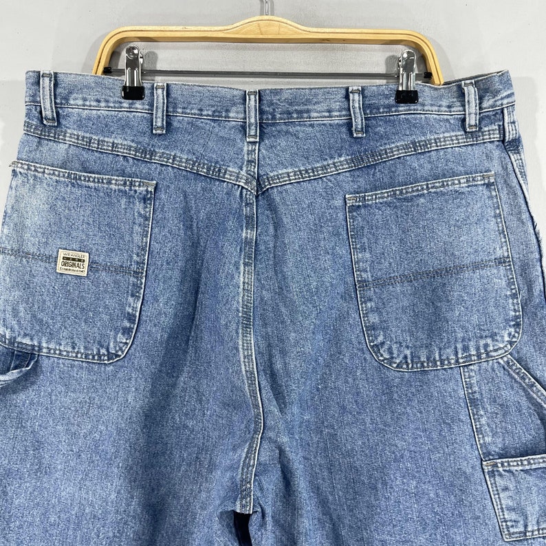 K&ouml;nnte beinhalten: Eine blaue Jeanshose im Carpenter-Stil mit einer Ges&auml;&szlig;tasche, auf der ein wei&szlig;er Etikett mit dem Text "Los Angeles Originals" angebracht ist.