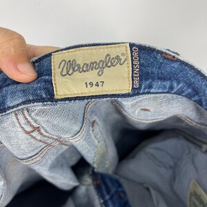 May include: Close-up of blue denim jeans with a Wrangler label. The label is tan with the Wrangler logo and "1947" printed on it. The word "GREENSBORO" is printed vertically on the right side of the label.
