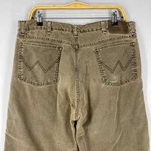 May include: Close-up of tan-colored denim jeans with the back pockets visible. The jeans have a classic five-pocket design, with the signature "W" stitching on the back pockets. A brown leather patch is visible at the waistband.
