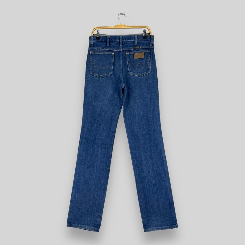 K&ouml;nnte beinhalten: Eine blaue Jeans aus Denim, die an einem Holzb&uuml;gel h&auml;ngt. Die Jeans hat einen klassischen geraden Schnitt und ein braunes Leder-Etikett mit dem Markennamen auf der R&uuml;ckseite.