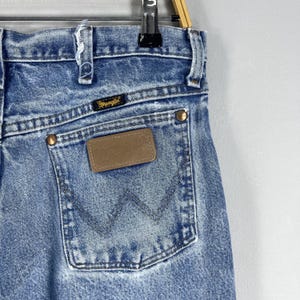 Pu&ograve; includere: Un paio di jeans blu denim con una toppa in pelle marrone che dice "Wrangler" sulla tasca posteriore.