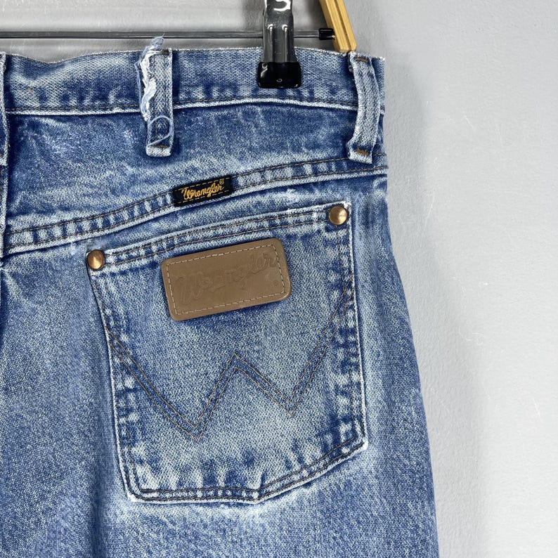 Pu&ograve; includere: Un paio di jeans blu denim con una toppa in pelle marrone che dice "Wrangler" sulla tasca posteriore.
