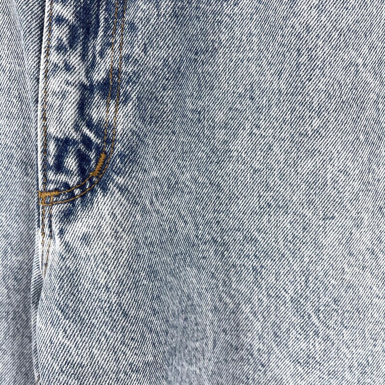 Pode incluir: Close-up de tecido de ganga azul claro com uma costura a correr verticalmente no lado esquerdo.