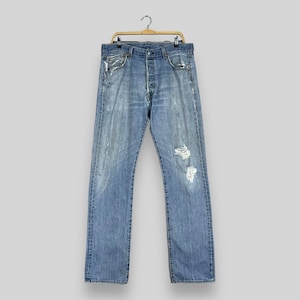 Puede incluir: Un par de pantalones vaqueros azules con detalles desgastados, colgados de una percha de madera con un gancho plateado.