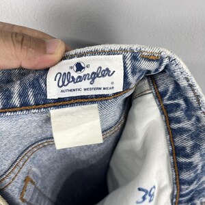 Pode incluir: Jeans de denim azul claro com uma etiqueta Wrangler. A etiqueta tem um logotipo de sino e o texto "Authentic Western Wear".