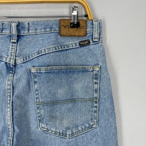 Puede incluir: Vaqueros de mezclilla azul claro con un bolsillo trasero y la marca "Wrangler" visible. Los vaqueros est&aacute;n colgados de una percha de metal.