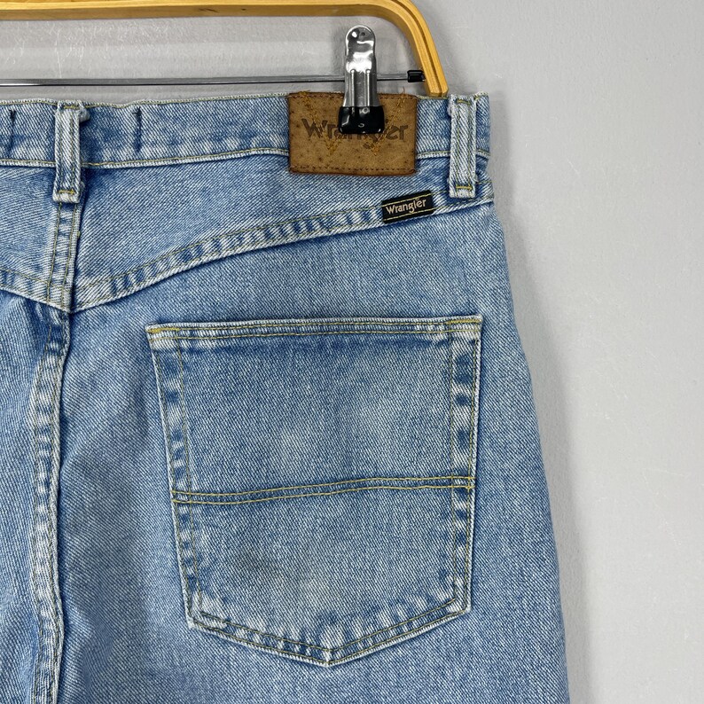 Puede incluir: Vaqueros de mezclilla azul claro con un bolsillo trasero y la marca "Wrangler" visible. Los vaqueros est&aacute;n colgados de una percha de metal.