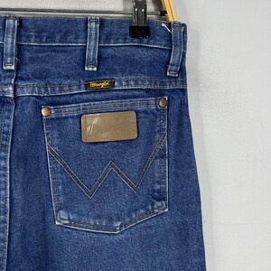 K&ouml;nnte beinhalten: Eine blaue Jeanshose mit einem braunen Leder-Patch auf der Ges&auml;&szlig;tasche, auf dem "Wrangler" steht.