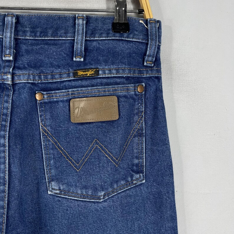 K&ouml;nnte beinhalten: Eine blaue Jeanshose mit einem braunen Leder-Patch auf der Ges&auml;&szlig;tasche, auf dem "Wrangler" steht.