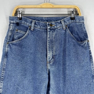 May include: A pair of blue denim jeans with a button fly and two front pockets. The jeans are hanging on a wooden hanger.