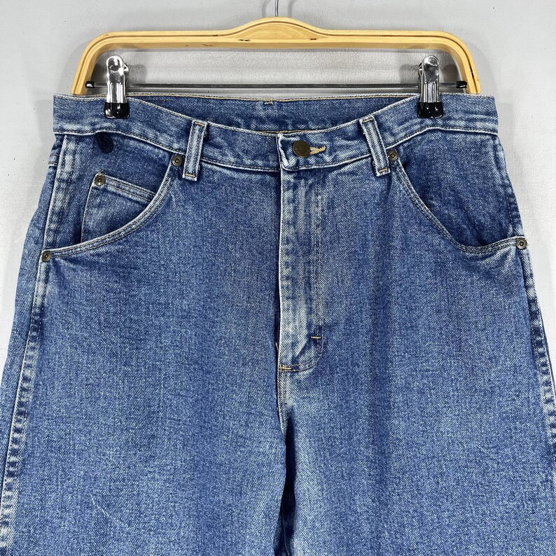 May include: A pair of blue denim jeans with a button fly and two front pockets. The jeans are hanging on a wooden hanger.