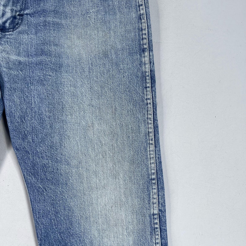 May include: Close-up of the side seam of a pair of light blue denim jeans. The seam is stitched with white thread.