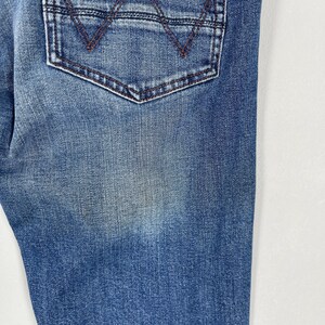 May include: Close-up of blue denim jeans with a back pocket. The pocket has a brown stitched design and a brown stitched border. The denim has a faded appearance.