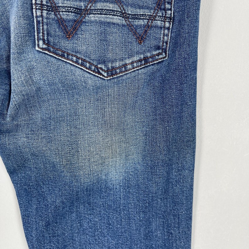May include: Close-up of blue denim jeans with a back pocket. The pocket has a brown stitched design and a brown stitched border. The denim has a faded appearance.