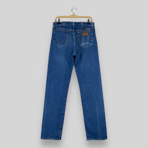 May include: A pair of blue denim jeans hanging on a wooden hanger. The jeans have a brown leather patch on the back pocket with the word "Wrangler" printed on it.