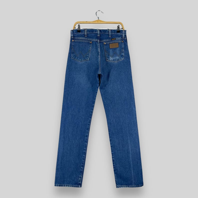 May include: A pair of blue denim jeans hanging on a wooden hanger. The jeans have a brown leather patch on the back pocket with the word "Wrangler" printed on it.