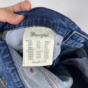 May include: Close-up of a Wrangler jeans label. The label is white with black text, listing the fabric composition as 99% cotton and 1% elastane. The brand name "Wrangler" is at the top. The label is attached to blue denim.