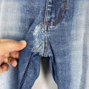 May include: Close-up of blue denim jeans with a distressed area near the crotch. The jeans have a faded wash and visible stitching in a contrasting brown color. The fabric is torn and frayed, showing the inner threads.