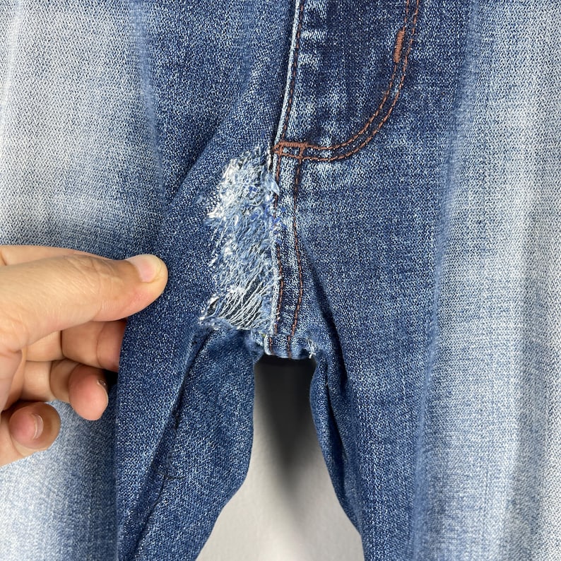 May include: Close-up of blue denim jeans with a distressed area near the crotch. The jeans have a faded wash and visible stitching in a contrasting brown color. The fabric is torn and frayed, showing the inner threads.