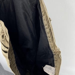 May include: Close-up of the interior of a pair of olive-green canvas pants. The inside is lined with black fabric. White stitching is visible along the seams and edges. A tag is visible at the bottom.