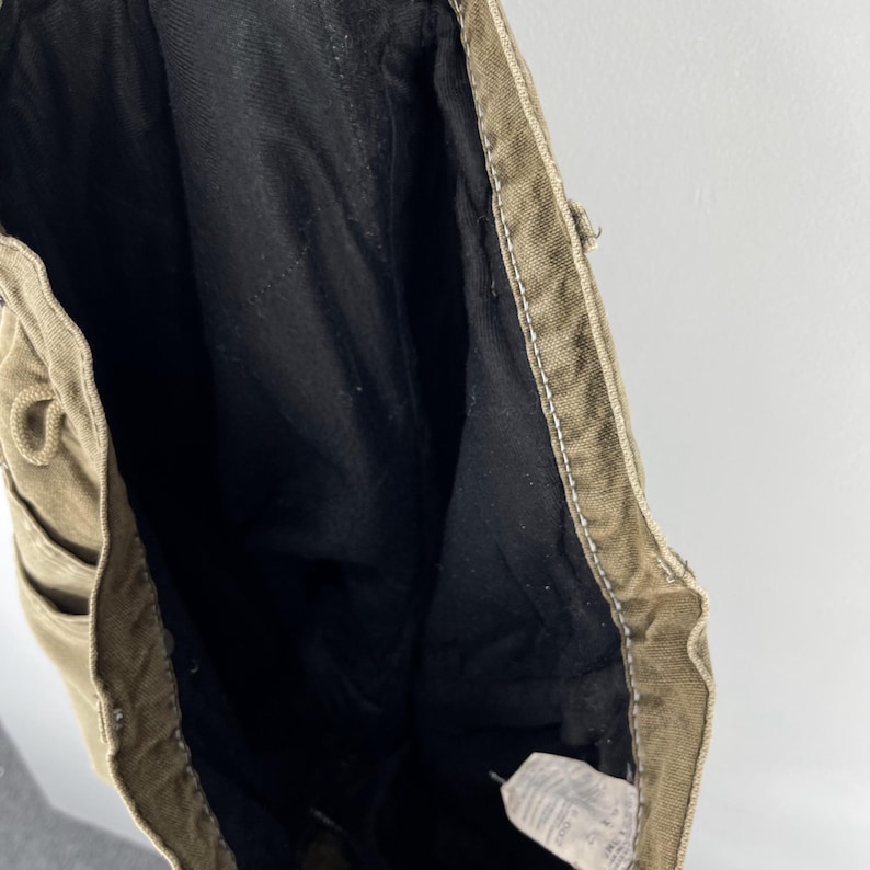 May include: Close-up of the interior of a pair of olive-green canvas pants. The inside is lined with black fabric. White stitching is visible along the seams and edges. A tag is visible at the bottom.