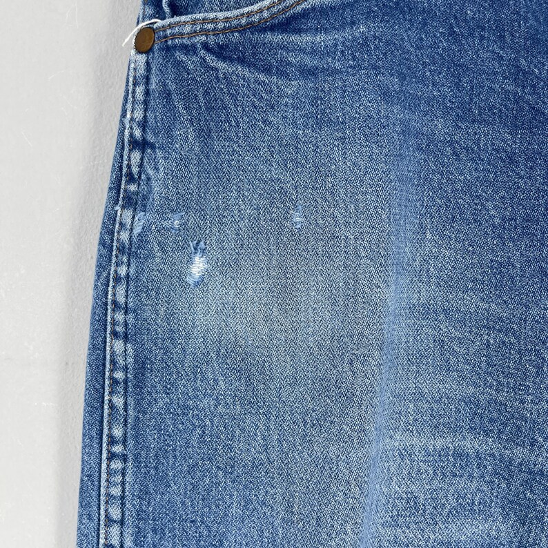 Pode incluir: Close-up de uma se&ccedil;&atilde;o de jeans azul. Os jeans mostram desgaste com &aacute;reas desbotadas e um pequeno rasgo. Um bot&atilde;o de lat&atilde;o &eacute; vis&iacute;vel na parte superior, junto com as costuras e uma parte do bolso.
