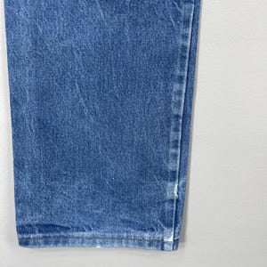 Pode incluir: Close-up de jeans de ganga azul. Os jeans t&ecirc;m um design cl&aacute;ssico de cinco bolsos e uma perna reta. O denim tem uma lavagem m&eacute;dia com algum desbotamento e desgaste. Os jeans s&atilde;o feitos de uma mistura de algod&atilde;o dur&aacute;vel.