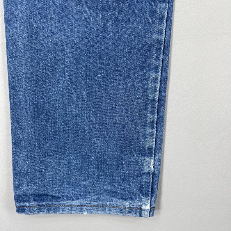 Pode incluir: Close-up de jeans de ganga azul. Os jeans t&ecirc;m um design cl&aacute;ssico de cinco bolsos e uma perna reta. O denim tem uma lavagem m&eacute;dia com algum desbotamento e desgaste. Os jeans s&atilde;o feitos de uma mistura de algod&atilde;o dur&aacute;vel.