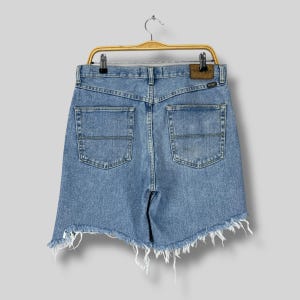 Puede incluir: Shorts vaqueros azul claro con dobladillo deshilachado, colgados de una percha de madera. Los shorts tienen dos bolsillos traseros y un parche de cuero marr&oacute;n con el nombre de la marca. Los shorts son de estilo vintage.