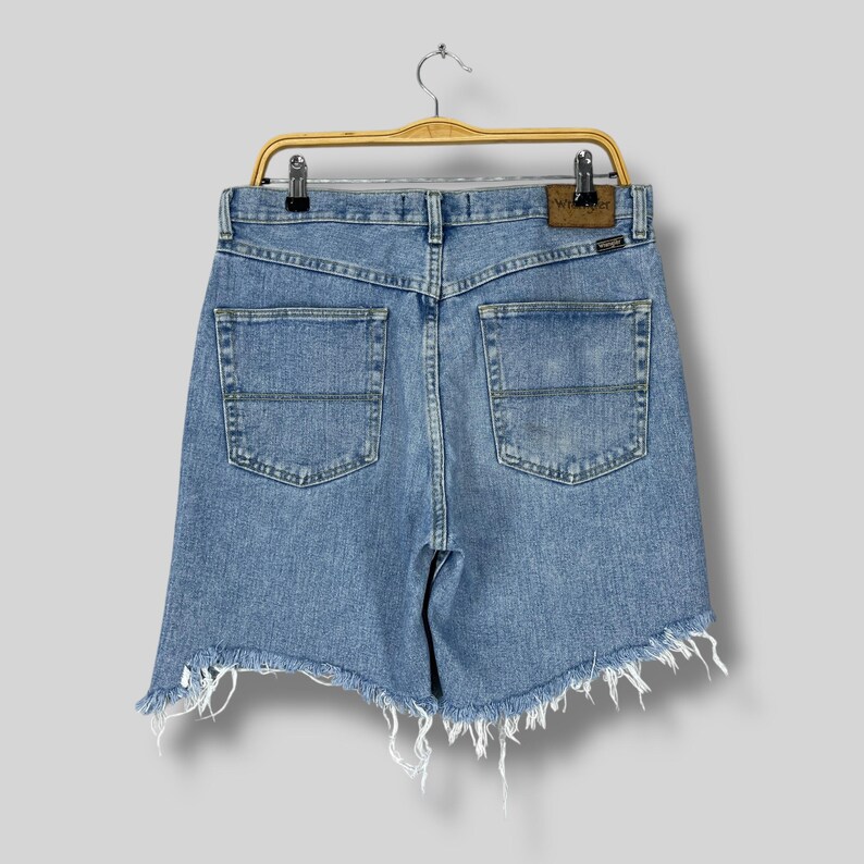 Puede incluir: Shorts vaqueros azul claro con dobladillo deshilachado, colgados de una percha de madera. Los shorts tienen dos bolsillos traseros y un parche de cuero marr&oacute;n con el nombre de la marca. Los shorts son de estilo vintage.