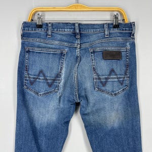 May include: Blue denim jeans with a classic five-pocket design. The back pockets feature a signature stitched 'W' design in a contrasting brown thread. A brown leather patch with the 'Wrangler' logo is visible.
