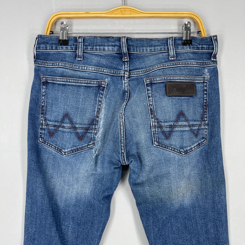 May include: Blue denim jeans with a classic five-pocket design. The back pockets feature a signature stitched 'W' design in a contrasting brown thread. A brown leather patch with the 'Wrangler' logo is visible.