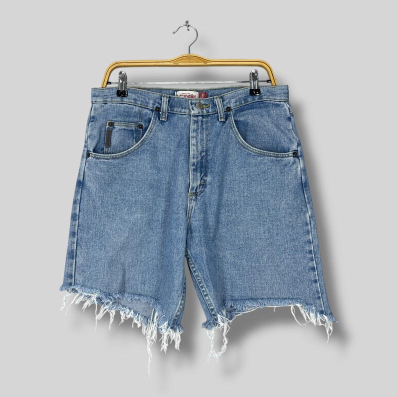 Puede incluir: Pantalones cortos vaqueros azul claro con dobladillo deshilachado, colgados de una percha de madera. Los pantalones cortos tienen un dise&ntilde;o cl&aacute;sico de cinco bolsillos y una etiqueta de la marca Wrangler. Los pantalones cortos son de lavado medio y parecen vintage.