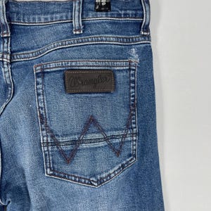 May include: Close-up of blue denim jeans with a Wrangler brand patch on the back pocket. The pocket has a stitched design in a darker blue thread. The jeans are faded and show signs of wear.