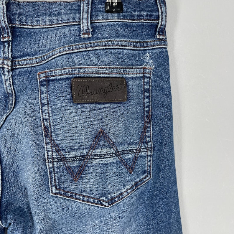 May include: Close-up of blue denim jeans with a Wrangler brand patch on the back pocket. The pocket has a stitched design in a darker blue thread. The jeans are faded and show signs of wear.