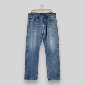 Puede incluir: Vaqueros de mezclilla azul claro con bragueta de botones, colgados de una percha dorada. Los vaqueros muestran signos de desgaste, con zonas descoloridas y pequeñas marcas de pintura. Un estilo clásico e informal.
