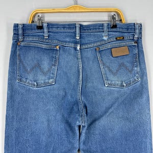 Pode incluir: Cal&ccedil;as de ganga azuis com um design cl&aacute;ssico de cinco bolsos. As cal&ccedil;as de ganga apresentam o log&oacute;tipo Wrangler num remendo de couro castanho e a costura W nas costas dos bolsos. As cal&ccedil;as de ganga est&atilde;o penduradas num cabide amarelo.