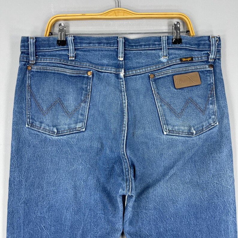 Pode incluir: Cal&ccedil;as de ganga azuis com um design cl&aacute;ssico de cinco bolsos. As cal&ccedil;as de ganga apresentam o log&oacute;tipo Wrangler num remendo de couro castanho e a costura W nas costas dos bolsos. As cal&ccedil;as de ganga est&atilde;o penduradas num cabide amarelo.